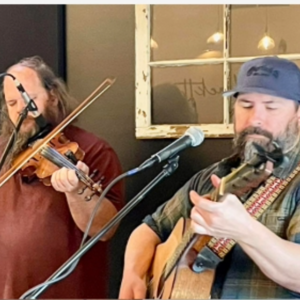 Two men performing live: a violinist on the left and a guitarist singing into a microphone on the right, both focused on their instruments.