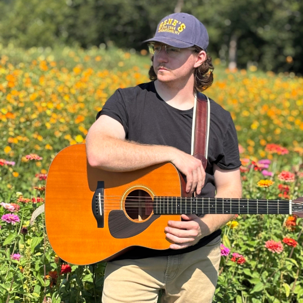 Person wearing a blue cap and black t-shirt holds an acoustic guitar outdoors in a vibrant flower garden.