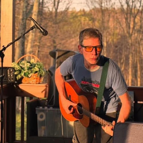 Man playing an acoustic guitar on an outdoor stage, wearing sunglasses and a blue T-shirt.