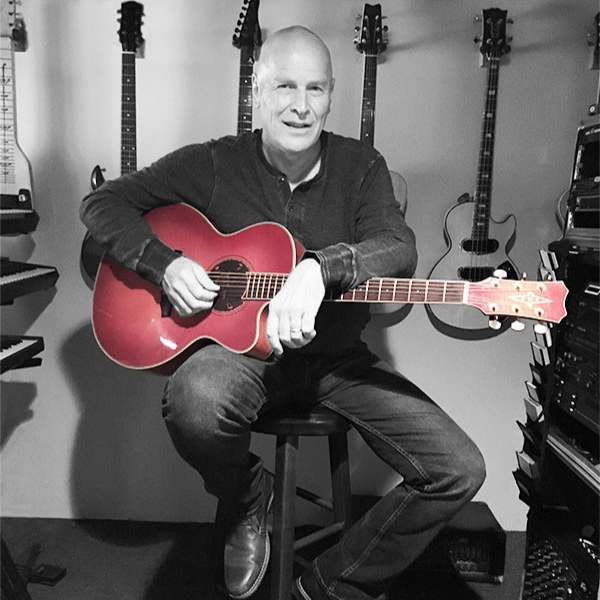 Man sits on a stool in a music studio, playing a red acoustic guitar with several guitars on the wall behind him in the background, color emphasized on the guitar.