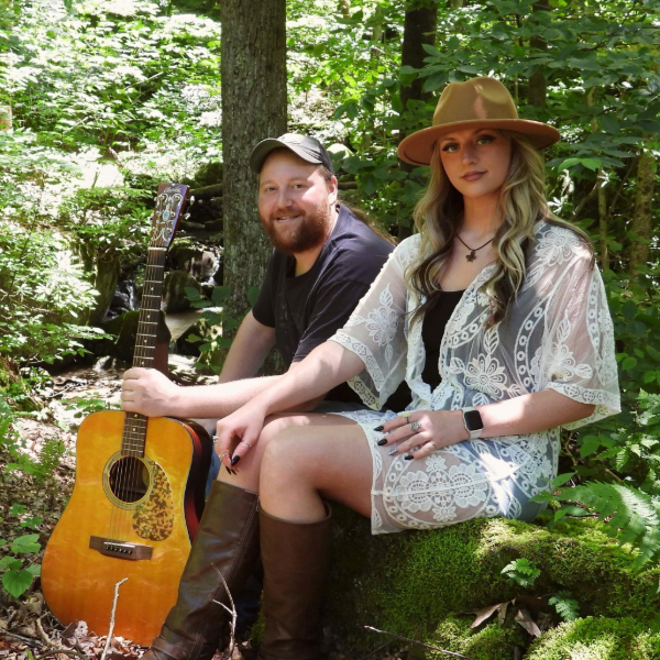 Two friends sit in a sunlit forest with an acoustic guitar beside them, suggesting a music duo outdoors.