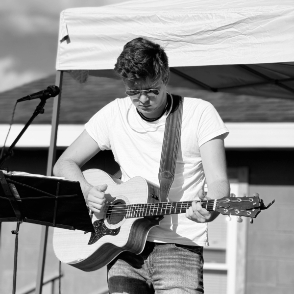 Male guitarist in a white t-shirt and sunglasses plays an acoustic guitar under a canopy at an outdoor performance.
