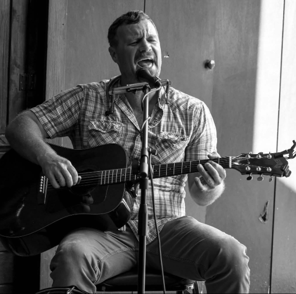 Male musician in a plaid shirt plays an acoustic guitar and sings into a microphone on a small stage.