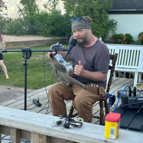 Bearded man in a bandana playing a resonator guitar outdoors, giving a thumbs-up toward the camera with a microphone nearby on a stand.